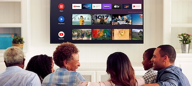 משפחה וחברים יושבים על ספה כאשר ב-Android TV שתלויה על הקיר מופיע תפריט בידור
