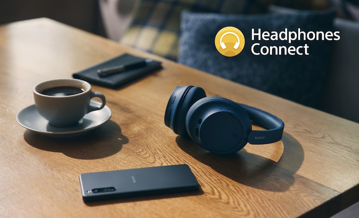 תמונה של אוזניות WH-CH720N כחולות על שולחן עם טלפון חכם, פנקס וכוס קפה. בפינה הימנית העליונה של התמונה מופיע לוגו של Headphones Connect.