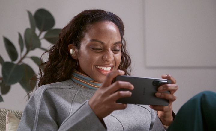 אישה יושבת בסלון ומרכיבה אוזניות LinkBuds S, צופה בסרטון בטלפון החכם שלה