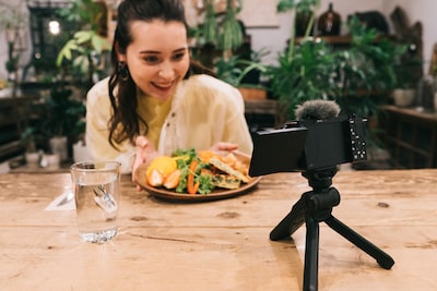 תמונה של אישה עם צלחת אוכל ליד שולחן, יושבת מול מצלמה שהוצבה על חצובה קטנה