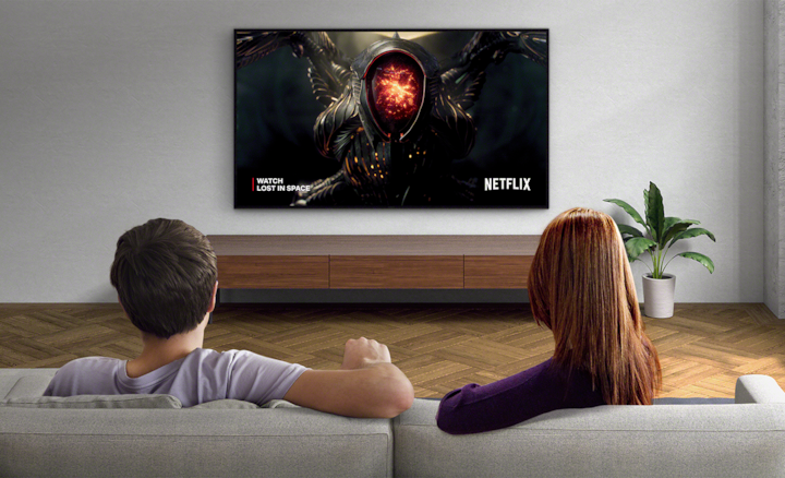 שני אנשים יושבים על ספה וצופים בטלוויזיה שמותקנת על קיר, עם צילום מסך של Lost in Space והלוגו של Netflix בצד ימין למטה