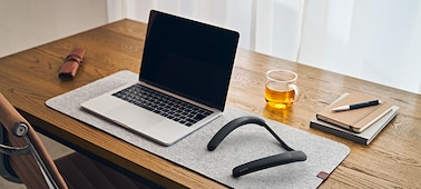 Objects on a wooden desktop, including notepads and a pen, an open laptop, a cup of tea, and the SRS-NB10 Wireless Neckband Speaker in black