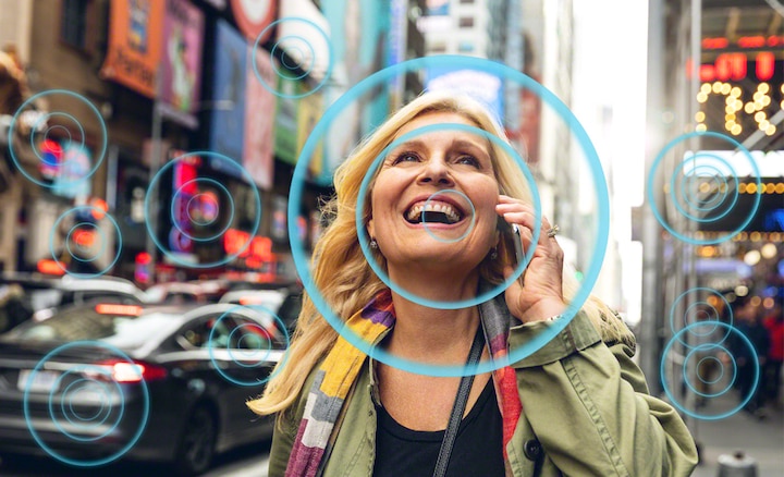 אישה ברחוב סואן מדברת בטלפון נייד, ומראה איך Voice Zoom 2 מקל על שמיעת דיבור