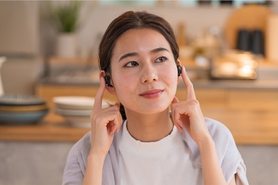 אישה יושבת במטבח ומאזינה לאוזניות WF-1000XM4 כשהאצבעות שלה מונחות על כל אוזניית כפתור