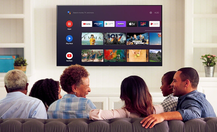 משפחה וחברים יושבים על ספה כאשר ב-Android TV שתלויה על הקיר מופיע תפריט בידור