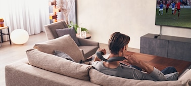 גבר יושב על ספה וצופה בסרט בטלוויזיה שמותקנת על הקיר בעזרת רמקול לביש SRS-NS7 ומשדר אלחוטי WLA-NS7