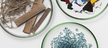 Overhead shot of three dishes containing different materials: wood and plant fibres, pieces of clear glass, and colourful plastic fragments