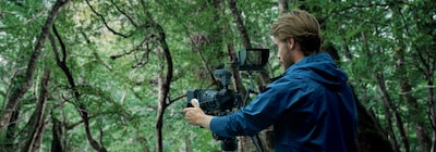 תמונה של גבר המשתמש במצלמה המחוברת למיקרופון חיצוני ולצג חיצוני, עם עלים ברקע