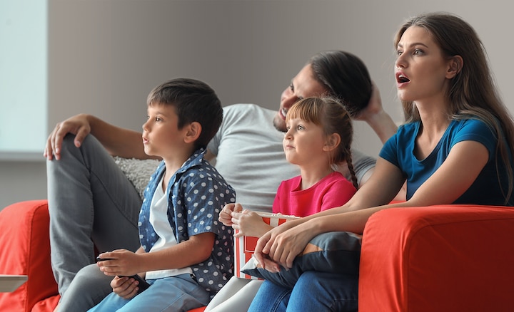 משפחה בת ארבע נפשות מנשנשת פופקורן על הספה האדומה בסלון וצופה במתח בטלוויזיה