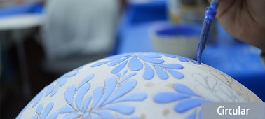 תמונה לדוגמה המציגה אומן מעטר צנצנת לבנה באמצעות מברשת וצבע כחול. המיקוד הוא במברשת, בעוד קדמת הצנצנת והרקע מטושטשים